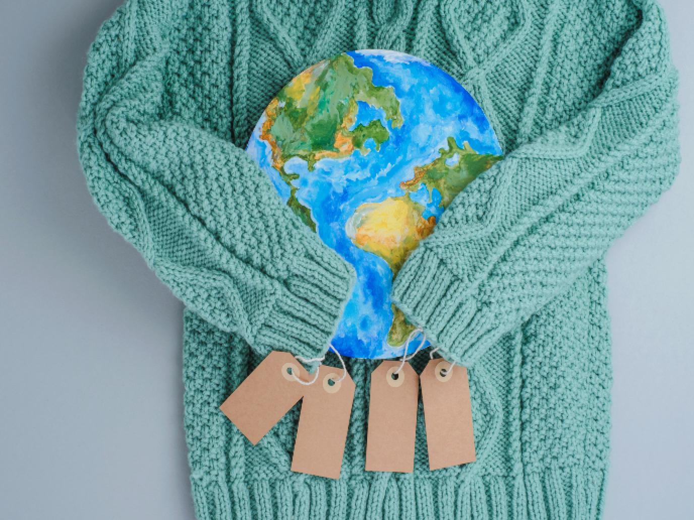 A green knit sweater with price tags attached to the arms holding a drawing of Earth